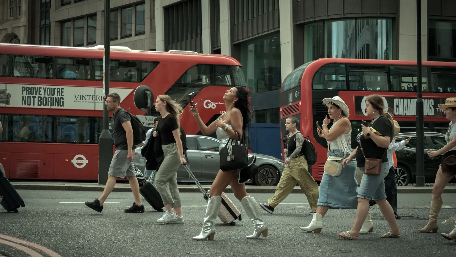 People crossing a London street with red buses passing behind